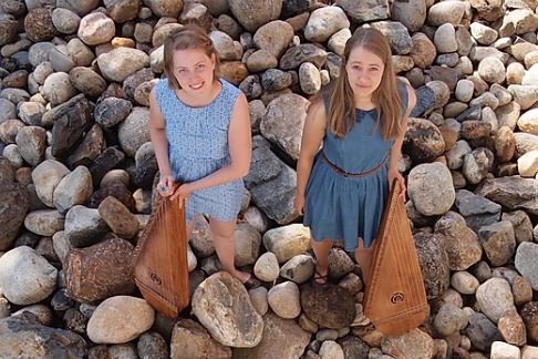 Tikkursby Duo, Kati Rantala & Jenni Venäläinen, kantele Kati och Jenni står på små stenar på en strand med sina tikkukantele i handen.