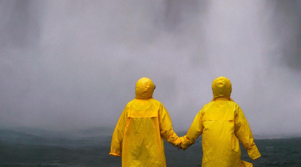 Oväder, två personer med gula regnrockar håller varandra i handen och ser ut mot ovädret.